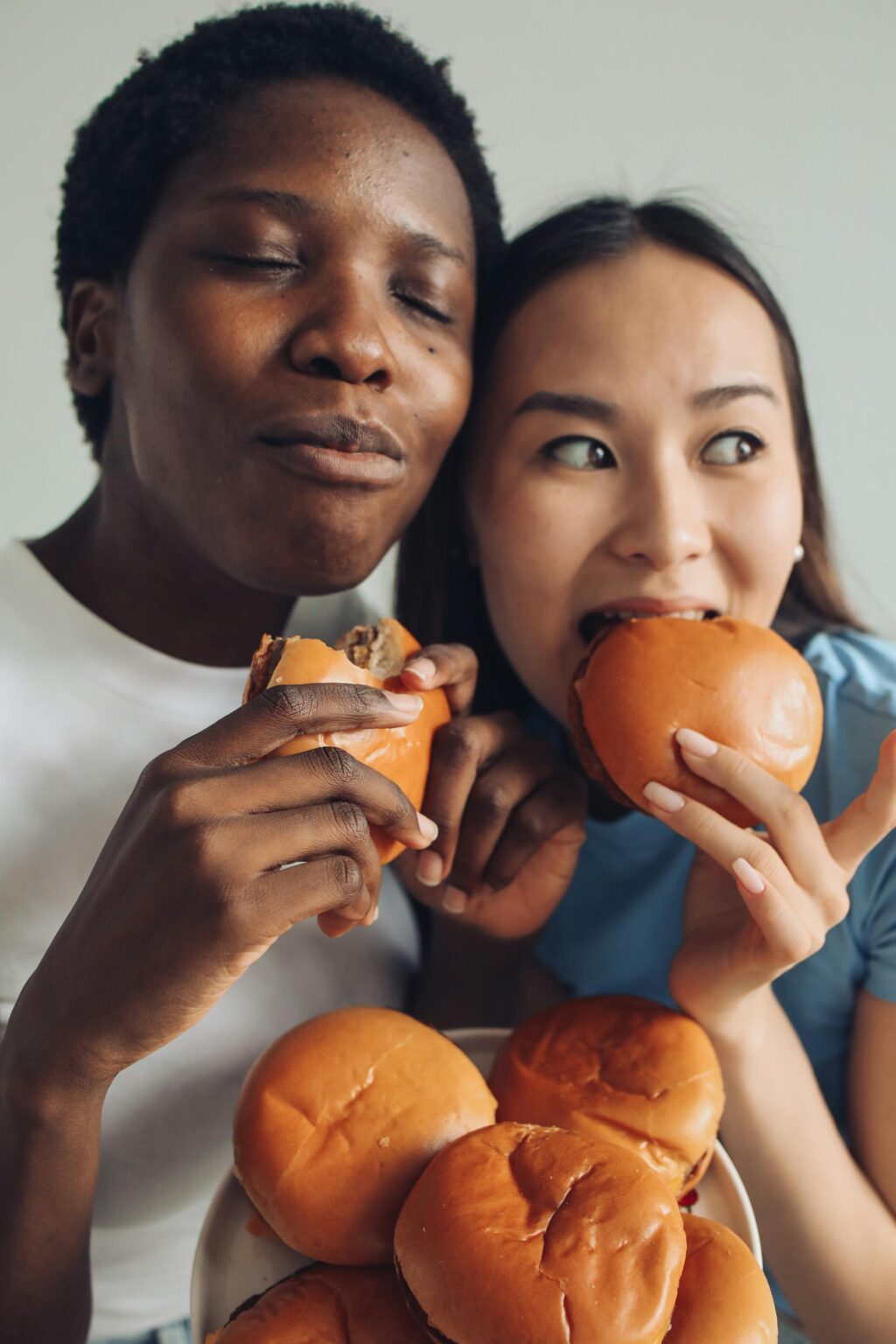Close-up of two friends eating burgers, sharing a joyful meal and laughter indoors.